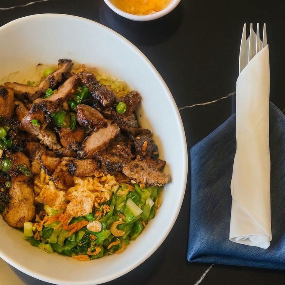 Bowl of grilled meat, mushrooms, rice, greens, and crispy toppings beside a rolled napkin on a blue cloth.